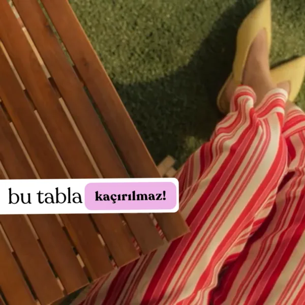 Teak ağacından yapılmış masif ahşap masa tablasının üstten çekilmiş görüntüsü. Masanın doğal ahşap çizgileri ve zarif dokusu dikkat çekiyor. Yan tarafta kırmızı beyaz çizgili bir elbise giymiş kişi, sarı ayakkabılarıyla görülüyor. Masanın altındaki yeşil çim arka planı doğal bir atmosfer yaratıyor.